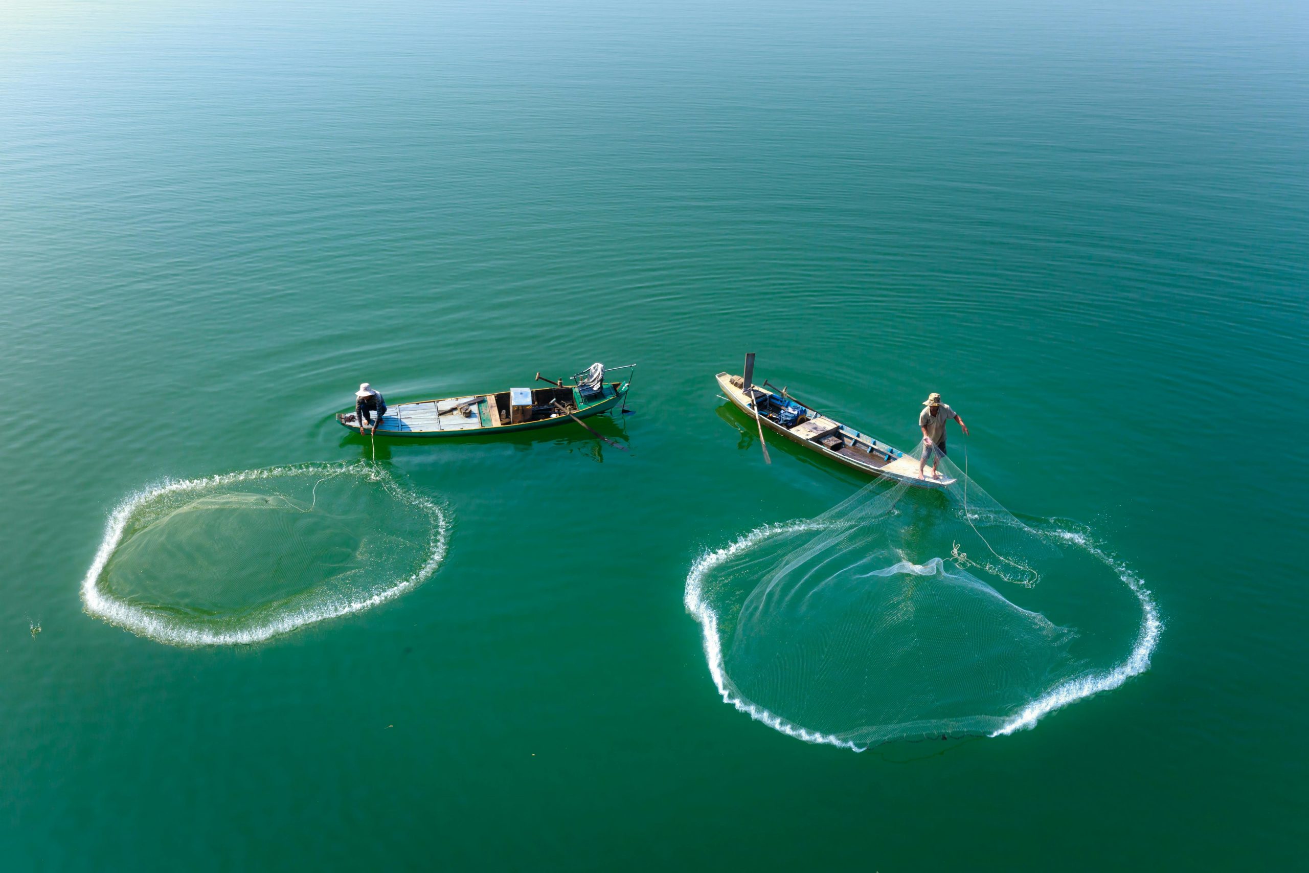 Drone shot capturing fishermen in boats casting nets on serene green water.