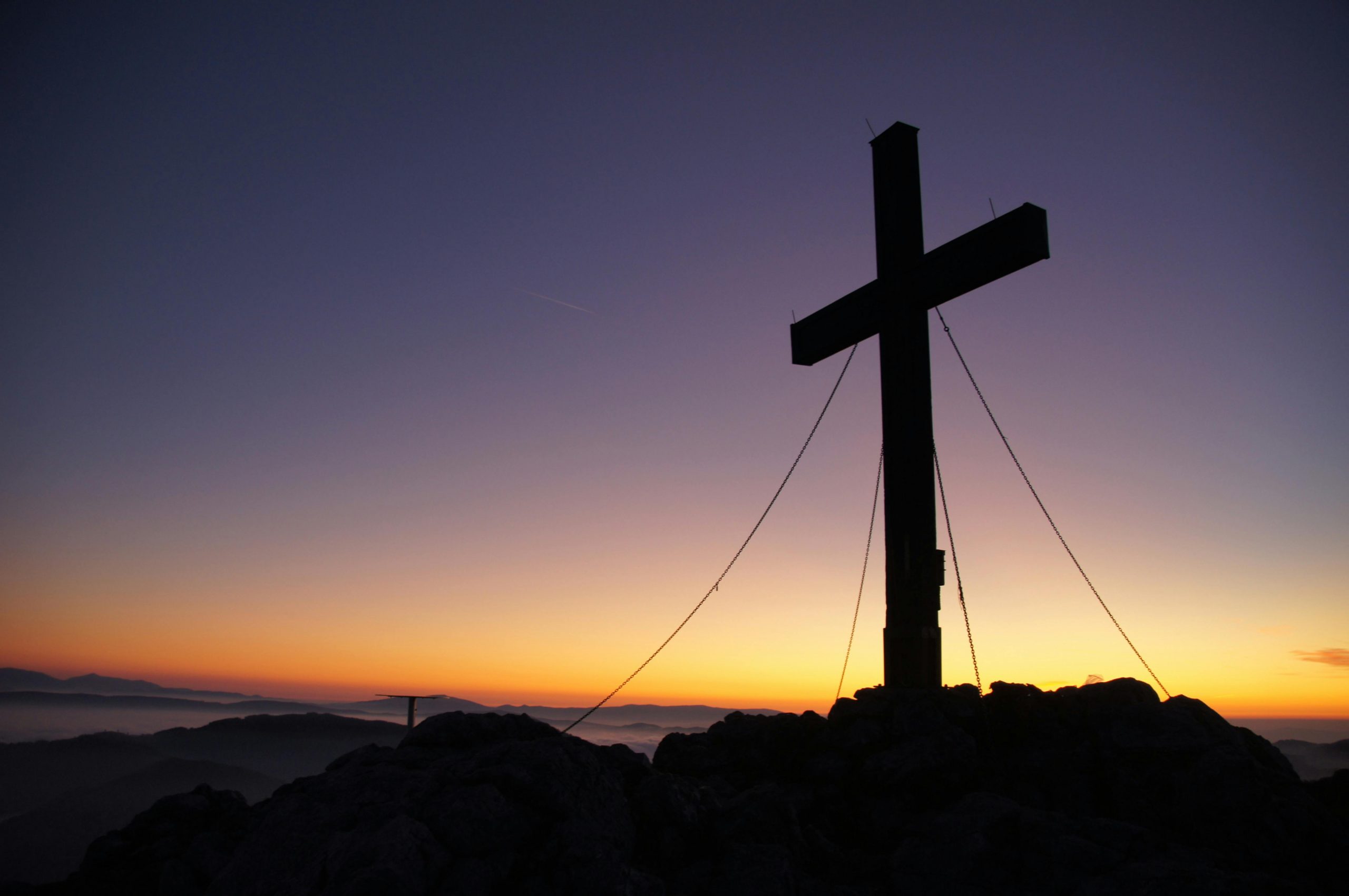 A dramatic cross stands silhouetted against a colorful sunset on a mountain peak, symbolizing faith and peace.
