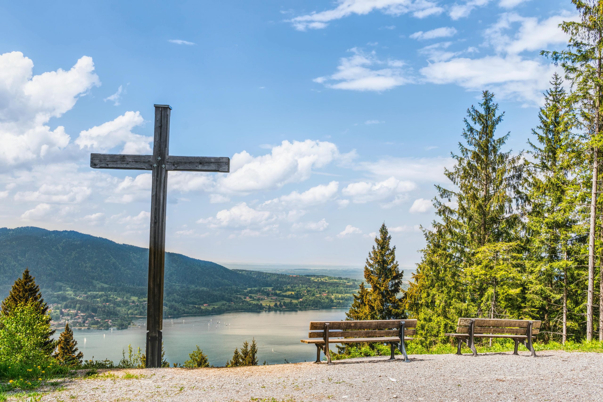 Serene landscape of Tegernsee in Germany featuring wooden cross, benches, and lush trees under a blue sky.