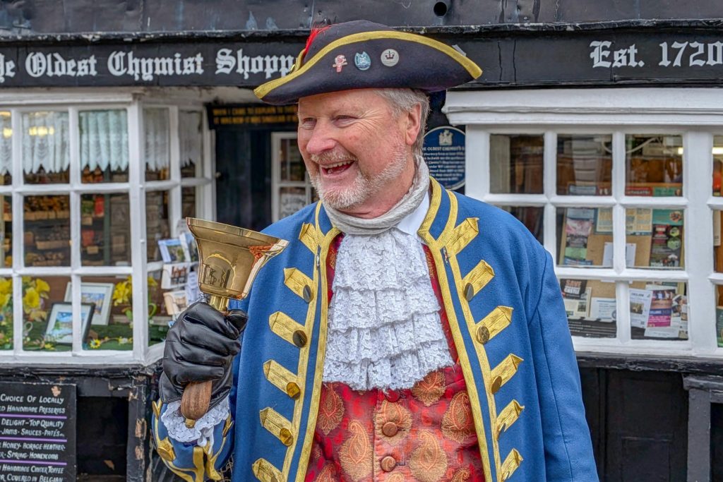 A cheerful town crier in traditional attire stands outside Ye Oldest Chymist Shoppe in England.