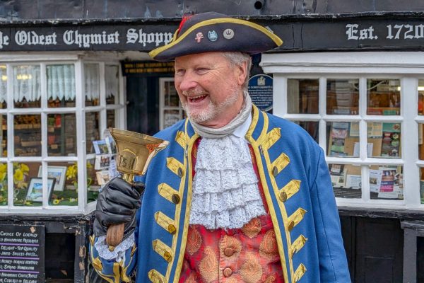 A cheerful town crier in traditional attire stands outside Ye Oldest Chymist Shoppe in England.