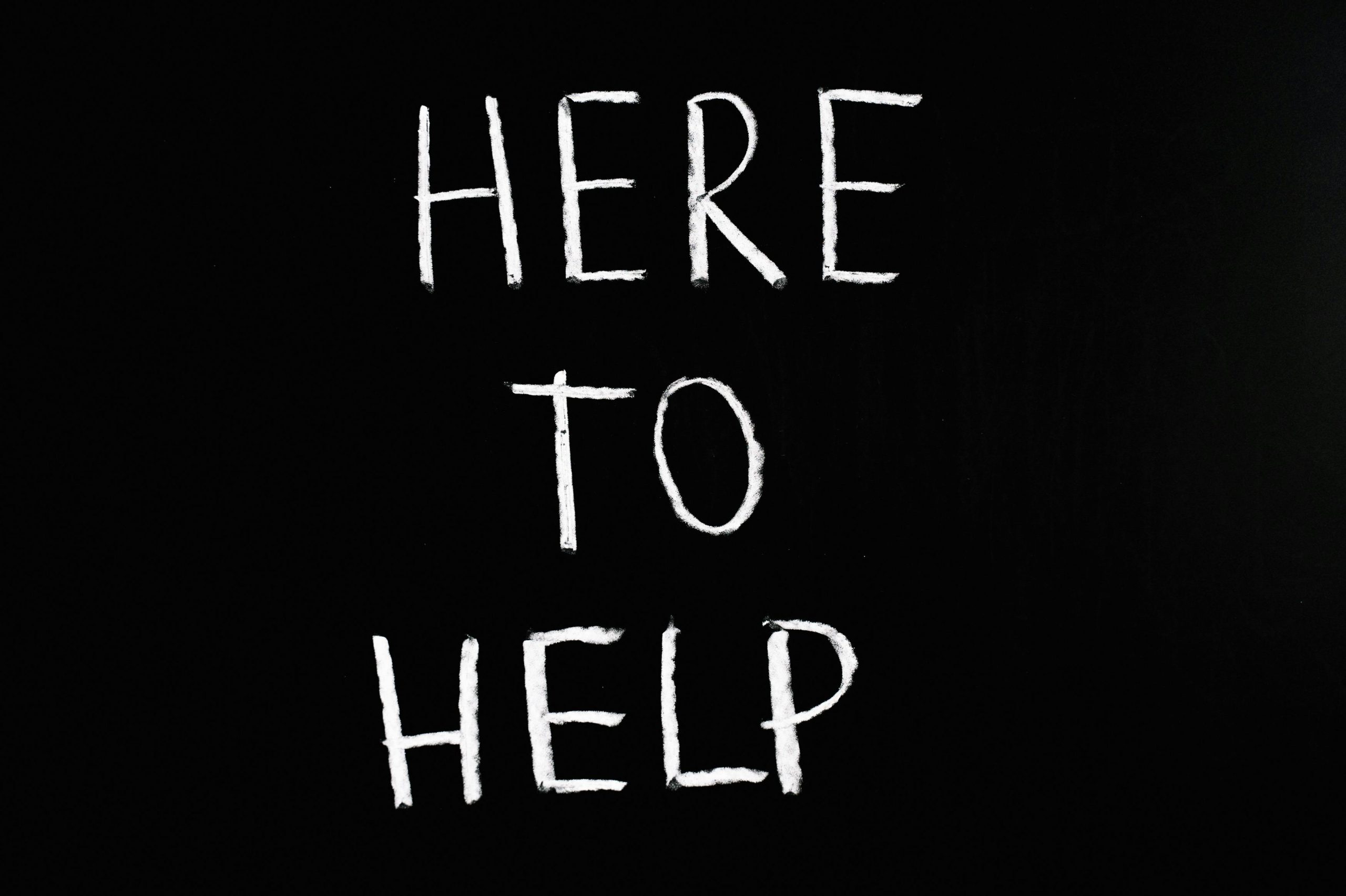 White text 'Here to Help' on a minimalist black chalkboard background, conveying encouragement and support.