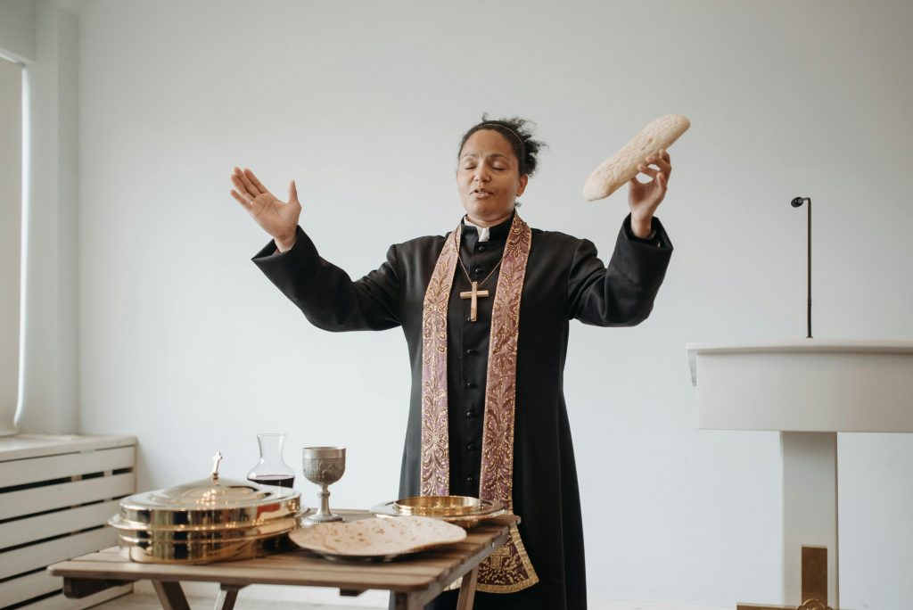 A female pastor leads a communion ceremony indoors, holding sacramental bread.
