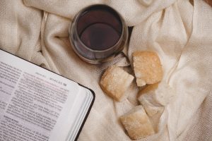 Overhead shot of a Bible, a glass of wine, and bread on a textured cloth.