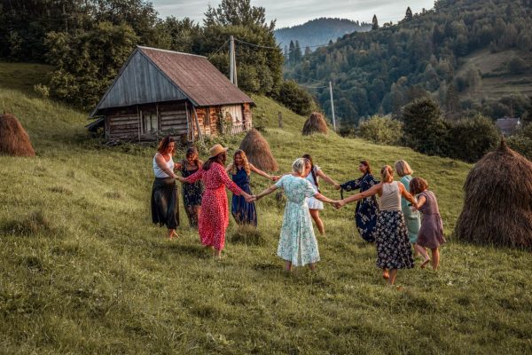 A group of women dancing in a rural meadow beside a wooden hut, surrounded by nature.