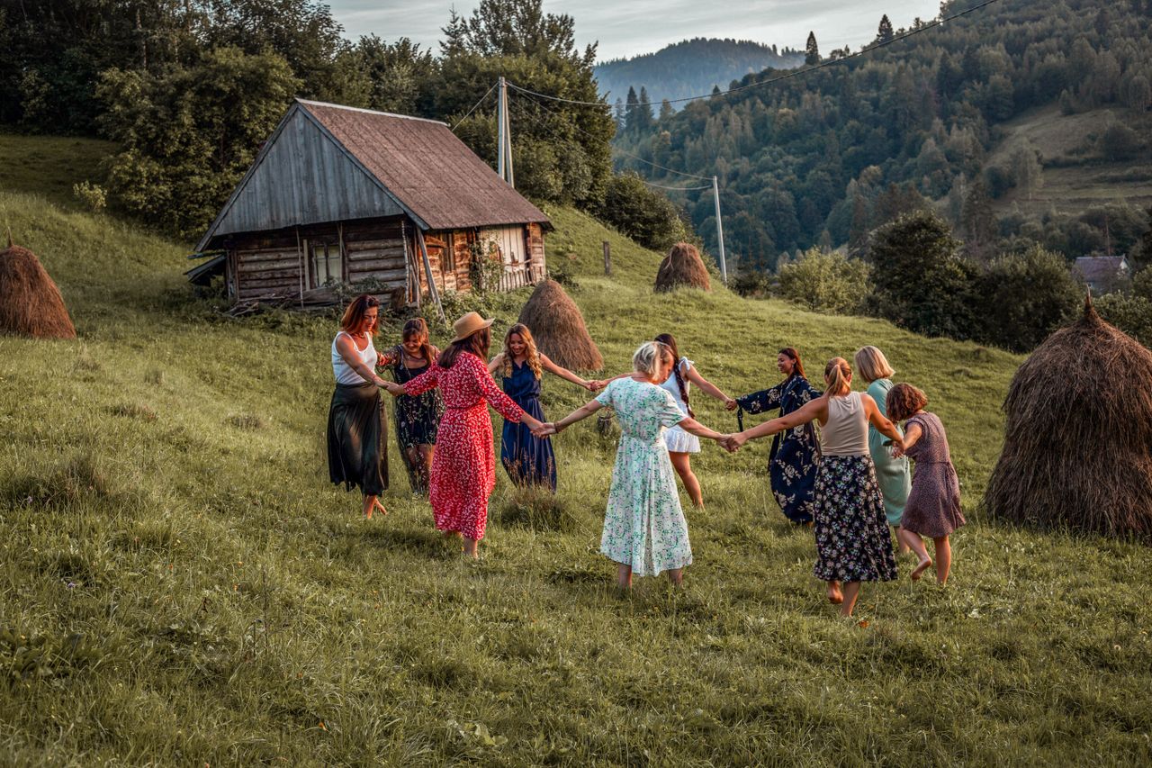 A group of women dancing in a rural meadow beside a wooden hut, surrounded by nature.